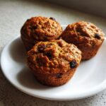 Three freshly baked, golden brown bran muffins studded with dark raisins resting on a white plate.