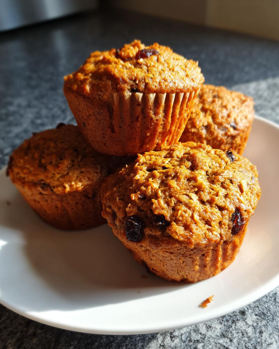 A stack of four golden brown, moist bran muffins dotted with raisins, resting on a white plate.
