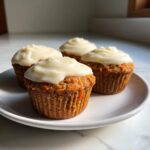 Four delicious carrot cake muffins topped with thick, white cream cheese frosting, resting on a white plate.