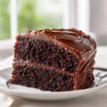 A perfect slice of moist chocolate cake layered with dark chocolate frosting, sitting on a white plate.