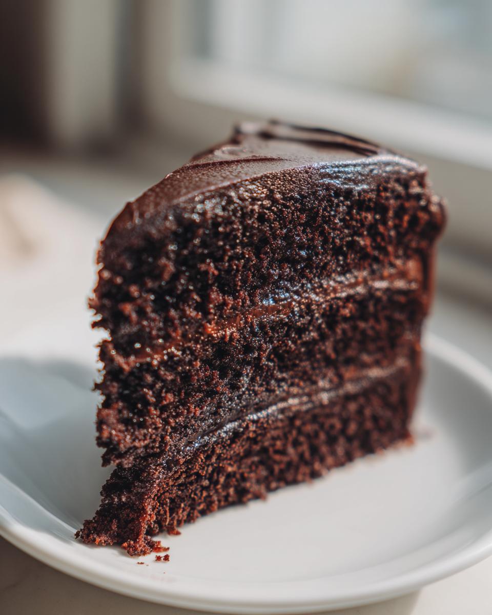 Close-up of a rich, moist chocolate cake slice layered with dark frosting on a white plate.