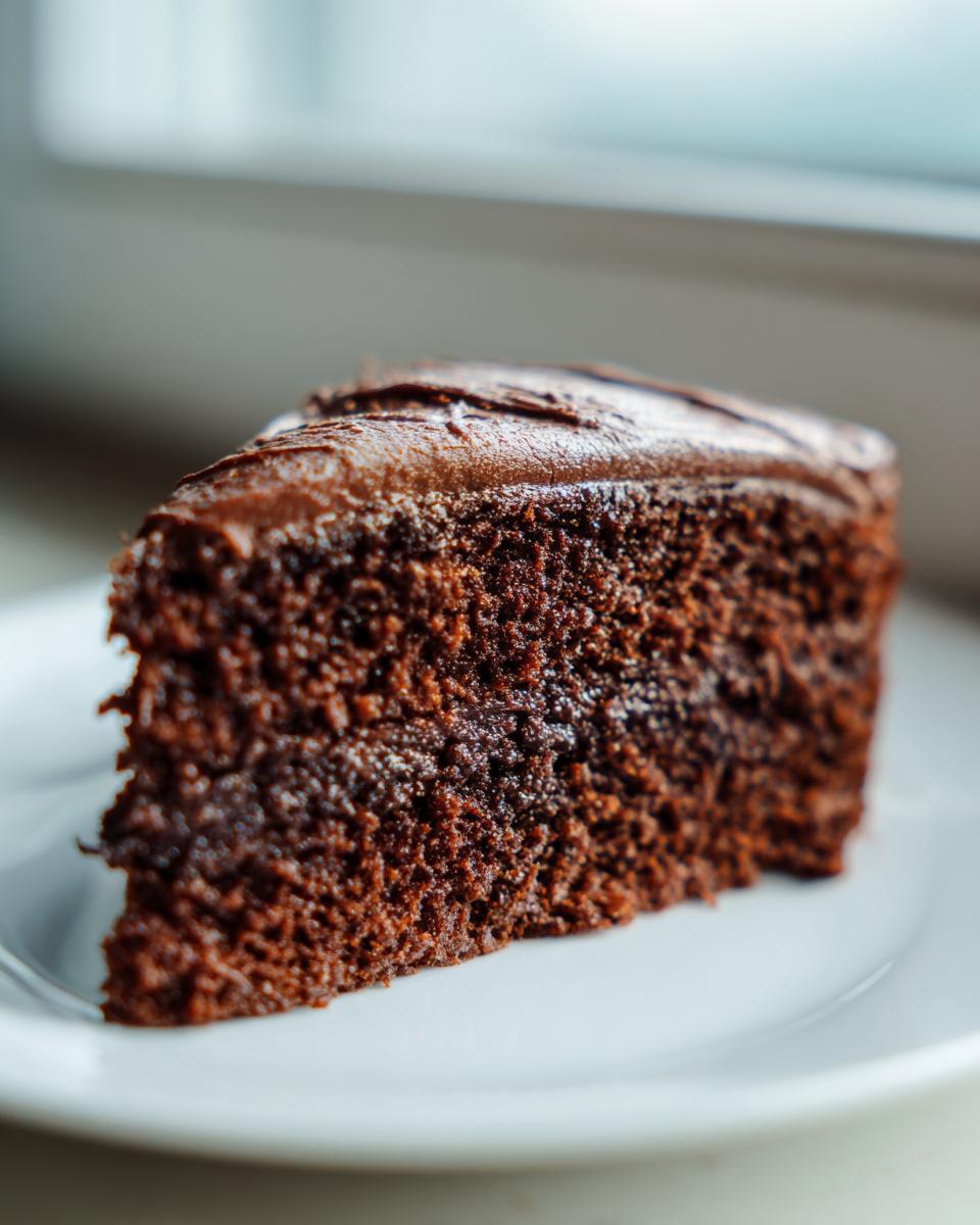 Close-up of a rich slice of moist chocolate cake topped with dark chocolate frosting on a white plate.