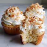 Three moist coconut cupcakes with white frosting and toasted coconut flakes, one has a bite taken out.