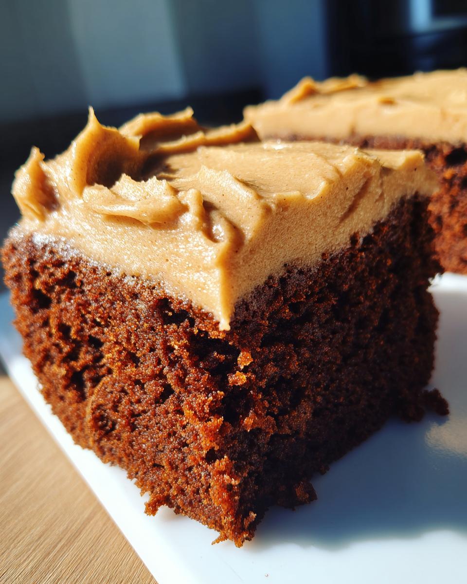 Close-up of a rich, dark slice of moist gingerbread cake topped with thick, light brown frosting.