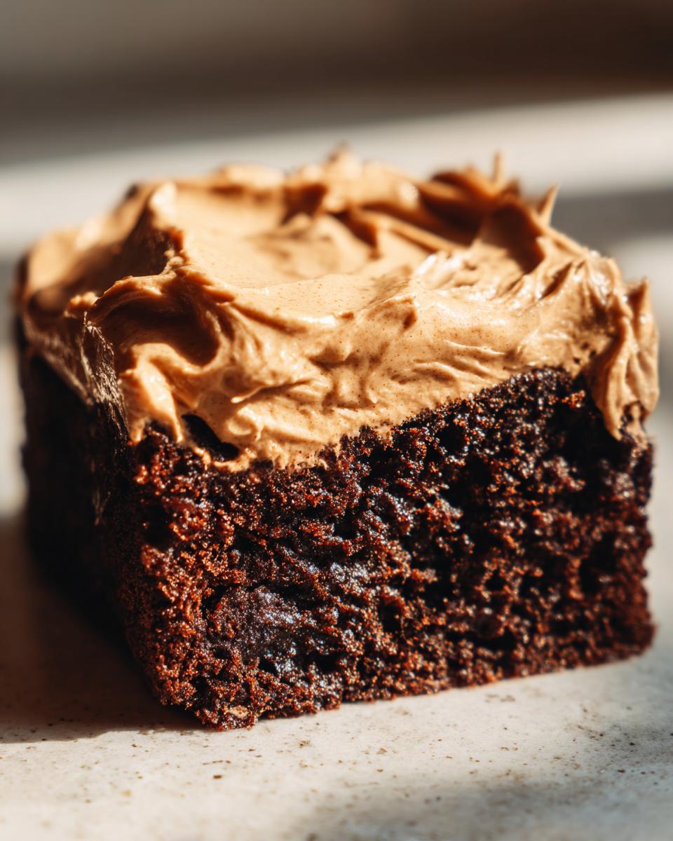Close-up of a moist, dark slice of gingerbread cake topped with thick, swirled brown frosting.