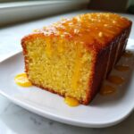 A close-up of a moist, yellow orange juice cake loaf drizzled with a thick, glossy orange glaze.