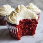Close-up of a moist red velvet cupcakes with a bite taken out, showing rich red crumb and thick cream cheese frosting.