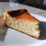 A close-up shot of a rich slice of New York cheesecake on a white plate, showing a dense, creamy interior and a deeply browned top.