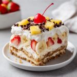 A decadent slice of no-bake banana split cake layered with cream, bananas, strawberries, pineapple, and topped with a cherry.