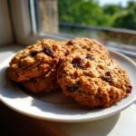 Three freshly baked oatmeal raisin cookies stacked slightly on a white plate near a sunlit window.