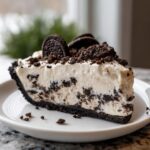 A thick slice of creamy Oreo pie with a dark crust, topped with crushed cookies and whole Oreos.