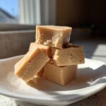 A stack of creamy, light brown peanut butter fudge squares resting on a white dish.