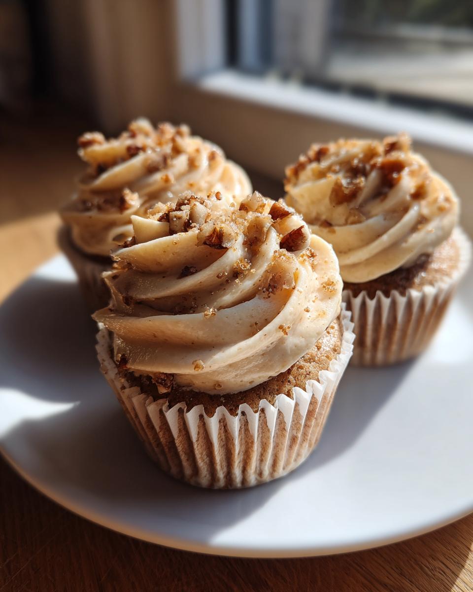 Three delicious pecan pie cupcakes topped with swirled brown frosting and chopped pecans, sitting on a white plate.