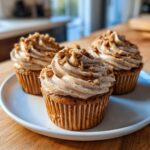 Three delicious pecan pie cupcakes with swirled, spiced frosting topped with chopped pecans, sitting on a white plate.