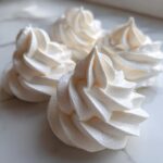 Close-up of four perfectly piped, white meringue cookies with crisp, swirled peaks resting on a marble surface.