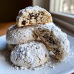 A stack of freshly baked cannoli cookies heavily dusted with powdered sugar, one cookie is broken open showing chocolate chips inside.