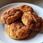 Close-up of four soft pumpkin snickerdoodle cookies coated heavily in cinnamon sugar, resting on a white plate.