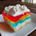 A square slice of colorful rainbow jello poke cake topped with swirls of white whipped cream.