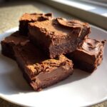 Close-up of rich, layered chocolate fudge squares stacked on a white plate, glistening in sunlight.