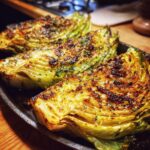 Three wedges of roasted cabbage recipe served on a dark platter, showing caramelized, browned tops sprinkled with herbs.