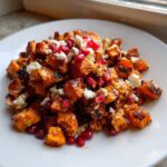 Close-up of roasted chunks in a sweet potato salad, topped with white feta cheese and bright red pomegranate seeds.