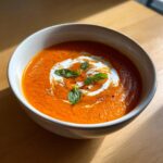 A vibrant bowl of roasted tomato soup topped with a swirl of cream, fresh basil leaves, and cracked black pepper.