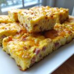 Close-up of stacked, golden-brown slices of sheet pan eggs loaded with ham and melted cheddar cheese.