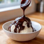 A spoonful of rich chocolate syrup is being drizzled over a scoop of vanilla ice cream in a white bowl.