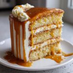 A tall slice of moist, three-layer caramel cake covered in white frosting and dripping with thick caramel sauce.
