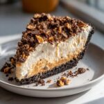 A close-up slice of rich butterfinger pie featuring a dark crust, peanut butter filling, whipped topping, and crushed Butterfinger candy.