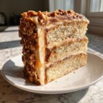 A tall slice of caramel crunch cake showing three layers of vanilla cake, caramel filling, and a thick coating of nuts.