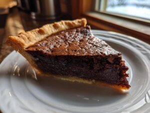 A close-up shot of a rich, dark slice of chocolate chess pie with a flaky crust on a white plate.