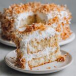 A tempting slice of layered coconut cake with white frosting and toasted coconut flakes sits on a small plate.