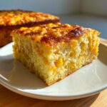 A square slice of moist cornbread casserole studded with whole corn kernels, sitting on a white plate.