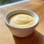 A small white bowl filled with creamy, pale yellow hollandaise sauce, sitting on a wooden surface.