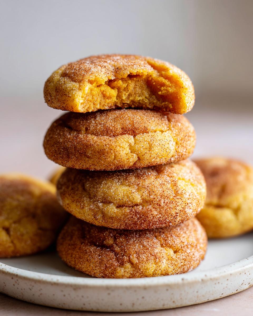 A stack of four soft pumpkin snickerdoodle cookies coated in cinnamon sugar, with the top one having a bite taken out showing the moist interior.