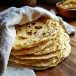 A stack of freshly made, warm corn tortillas wrapped partially in a light cloth on a wooden board.