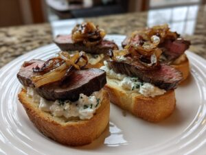Four pieces of steak crostini featuring medium-rare steak, creamy cheese spread, and caramelized onions on toasted bread.