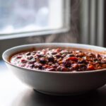 A close-up of a steaming white bowl filled with rich, dark black bean chili, set against a bright, blurred window background.