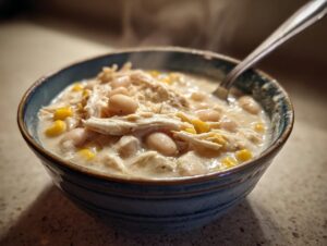 A close-up of a steaming bowl of creamy white cream cheese chicken chili with shredded chicken, white beans, and corn.