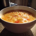 Close-up of a steaming white bowl filled with hearty cheese soup containing visible chunks of potato and carrot.