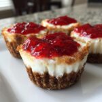 Four individual cheesecake bites with graham cracker crusts, creamy filling, and bright red strawberry topping.