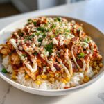 A close-up of a street corn chicken rice bowl topped with cotija cheese, crema, and spices.