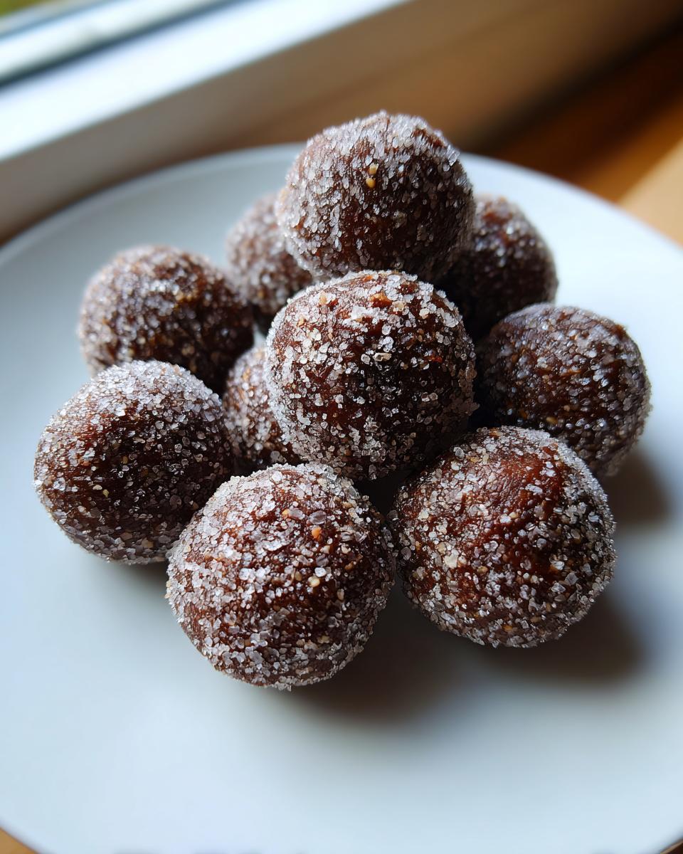 A close-up of several rich, dark bourbon balls heavily coated in sparkling granulated sugar, stacked on a white plate.