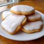 A stack of sugar cookies topped with thick, white sugar cookie icing and white sprinkles.