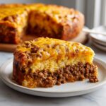 A thick slice of tamale pie showing a layer of seasoned ground meat topped with cornbread and melted cheddar cheese.