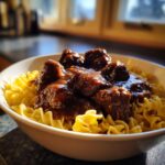 A close-up of tender crockpot beef tips smothered in rich brown gravy served over a bed of bright yellow egg noodles in a white bowl.