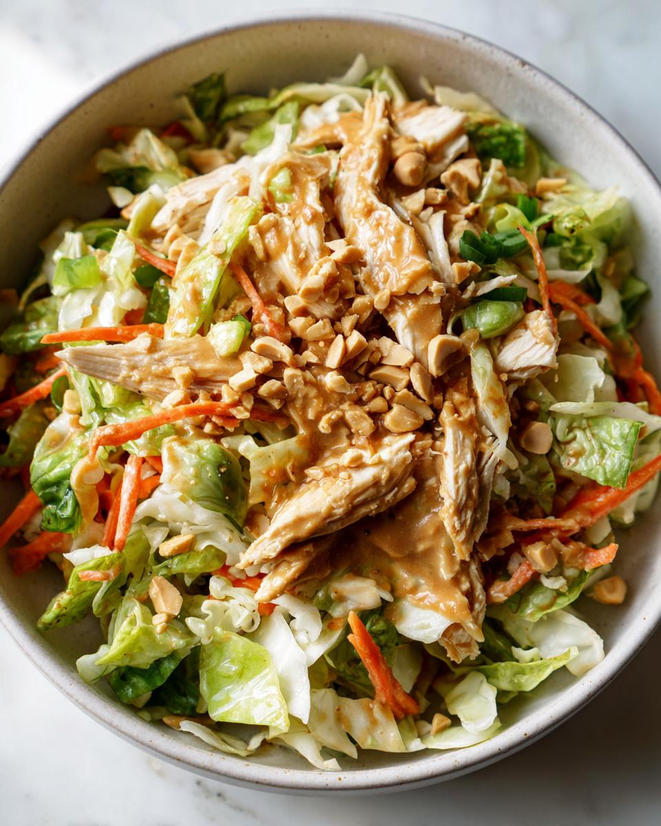 Close-up of a bowl of Thai chicken salad topped with shredded chicken, peanut dressing, and chopped peanuts.