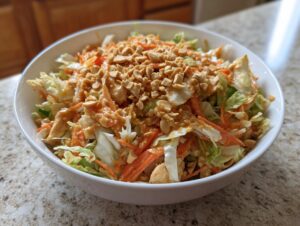 A white bowl filled with vibrant Thai chicken salad, topped generously with peanut dressing and chopped peanuts.