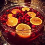 Close-up of a glass punch bowl filled with dark red Thanksgiving punch, garnished heavily with floating fresh cranberries and orange slices.